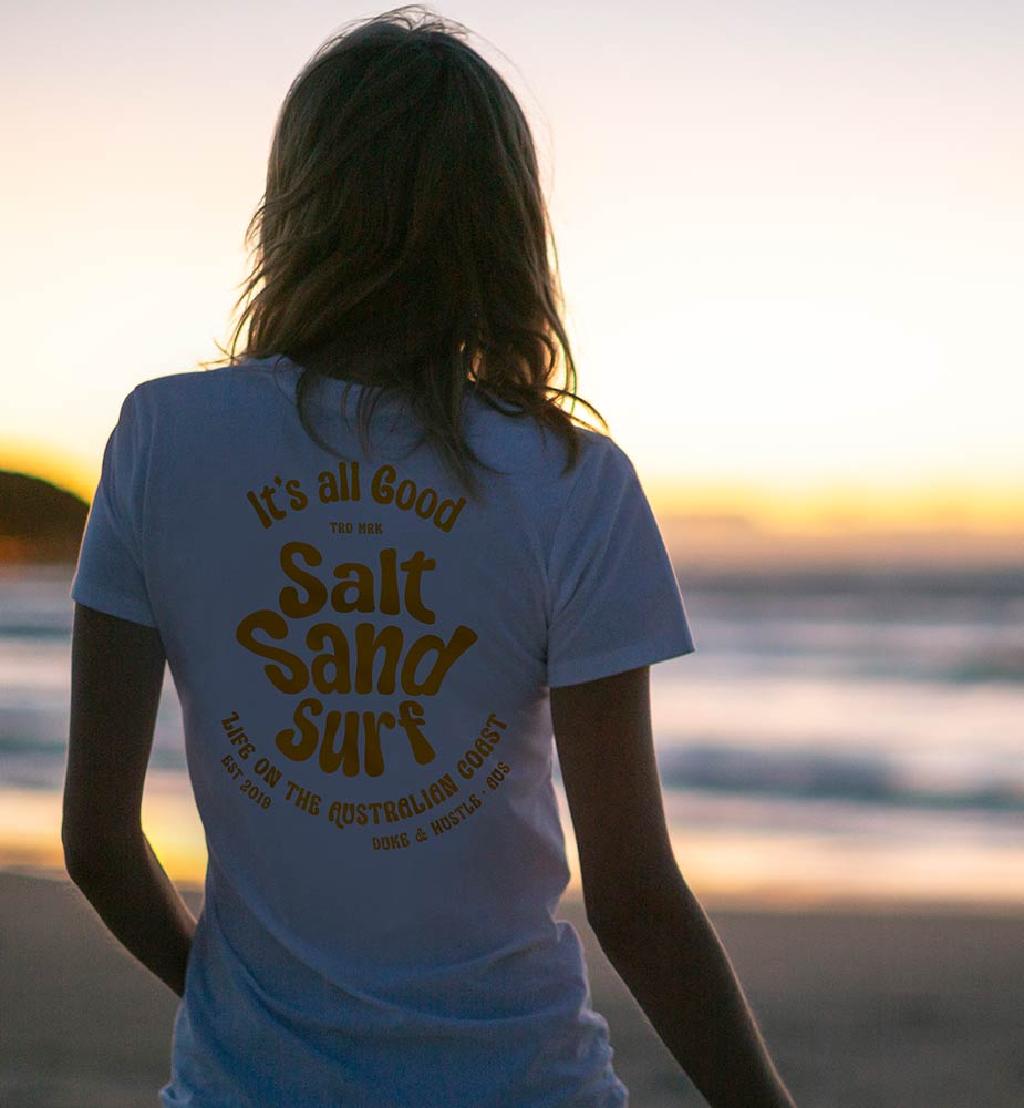 A woman looks out to the surf of a beautiful Australian beach at sunrise, wearing a Duke and Hustle cotton tee
